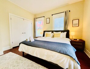 Ein Hotelzimmer mit Bett, Nachttisch, Lampe, einem Fenster mit Vorhängen, einer Tür und einem gerahmten Schild an der Wand.