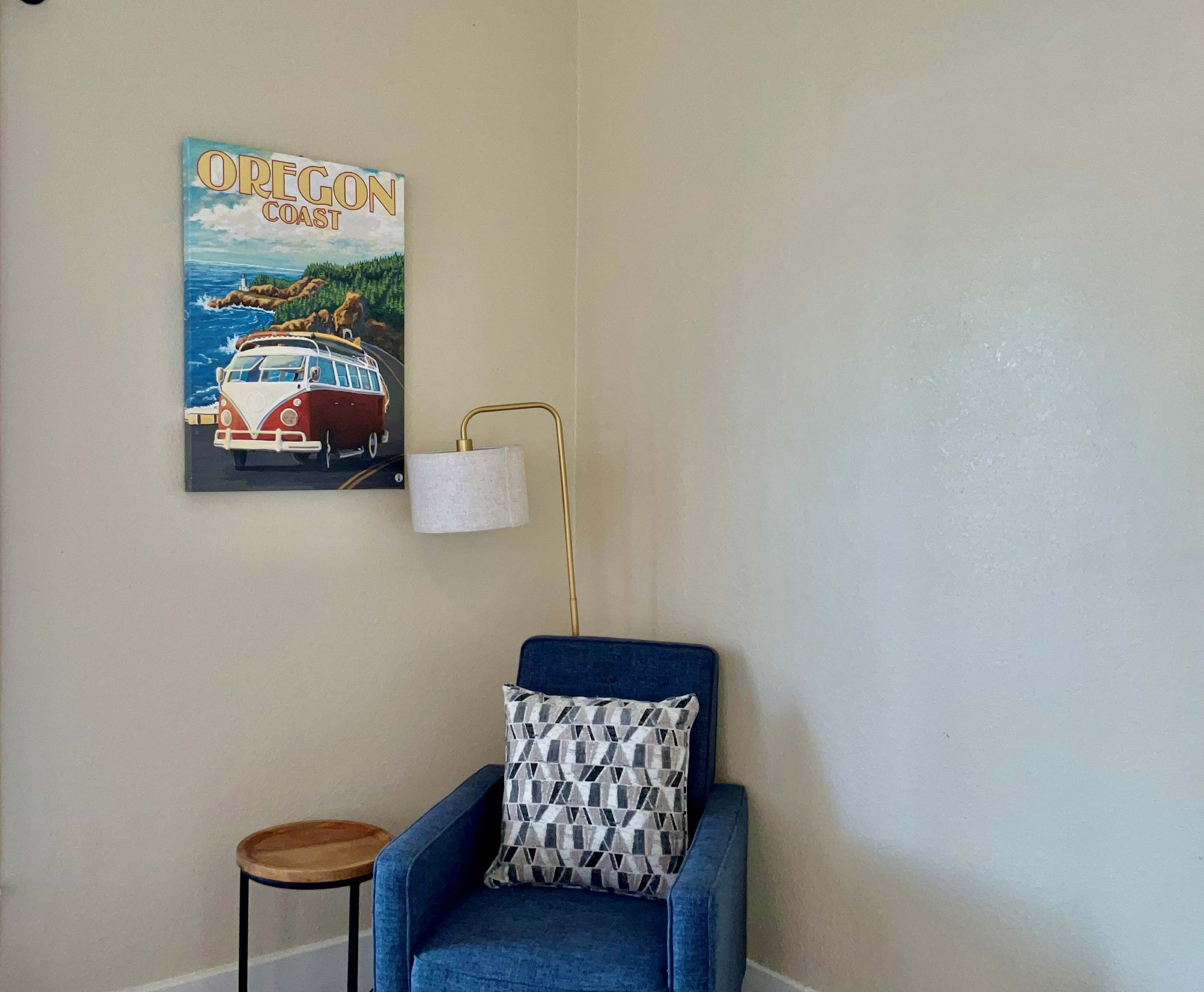 Een kamer met een blauwe fauteuil, een klein houten krukje, een wandlamp en een poster van een landschap aan de kust van Oregon.
