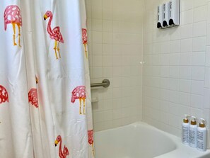 Un baño con cortina de ducha con diseño de flamencos, una bañera y tres botellas de artículos de tocador con las etiquetas 'champú', 'gel de ducha' y 'acondicionador'.