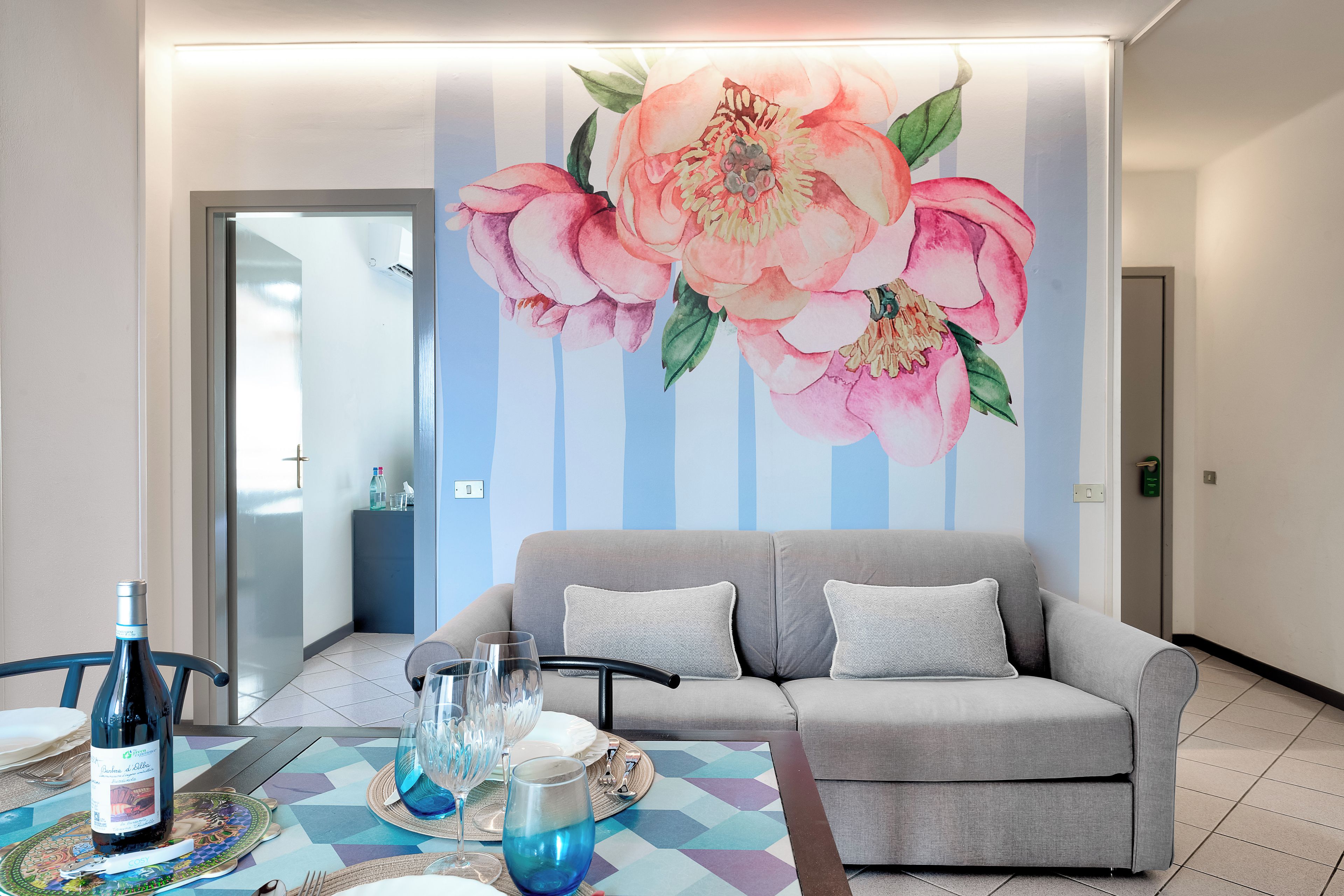 Una sala de estar moderna con un sofá gris, una mesa de comedor con platos, vasos y una botella, y un gran mural floral en la pared.