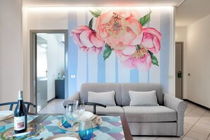 Una sala de estar moderna con un sofá gris, una mesa de comedor con platos, vasos y una botella, y un gran mural floral en la pared.