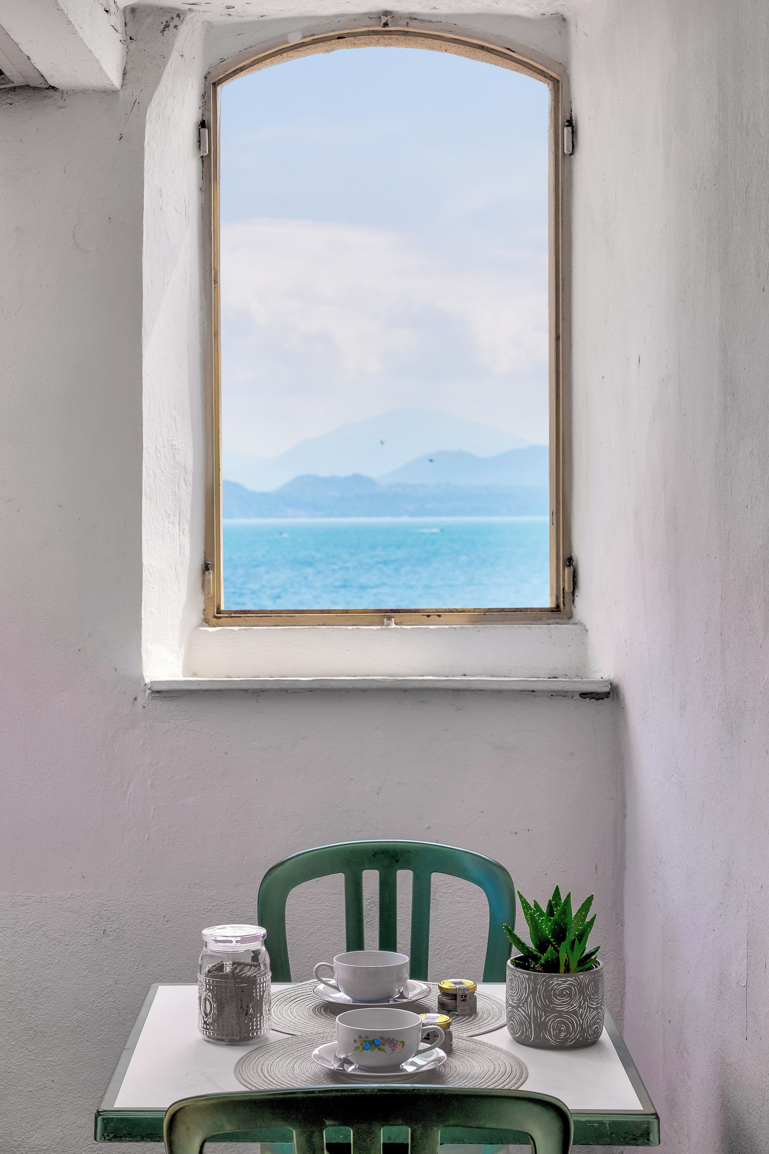 Une table placée devant une fenêtre donnant sur la mer, une chaise verte et une plante en pot.