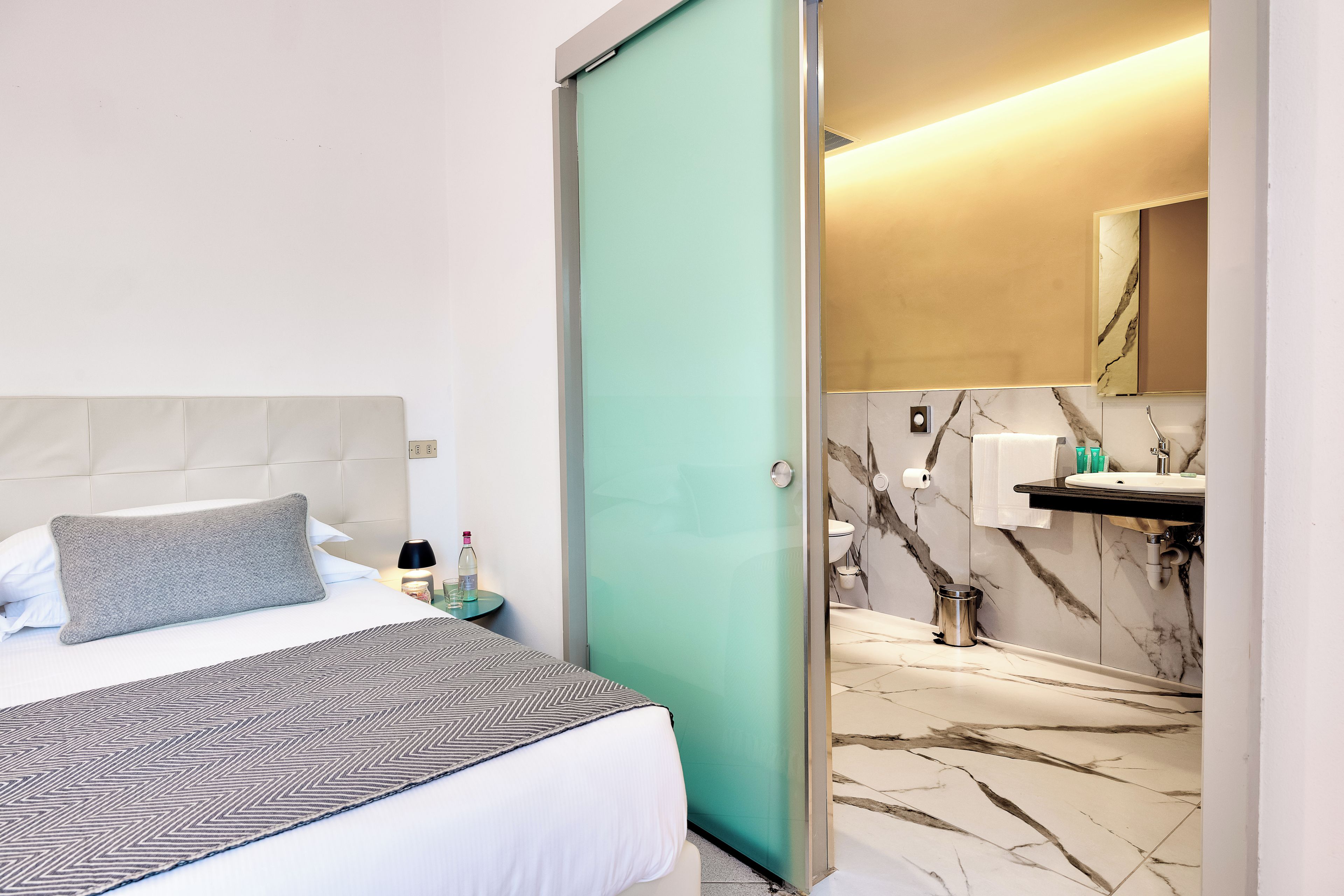 Een hotelkamer met een bed, nachtkastje en een badkamer met wastafel en spiegel.