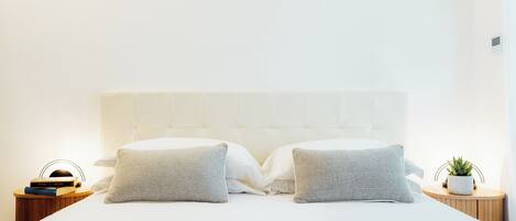 Cama bem arrumada com colcha estampada, dois travesseiros, mesa de cabeceira com um livro e uma planta, e termostato fixado na parede.