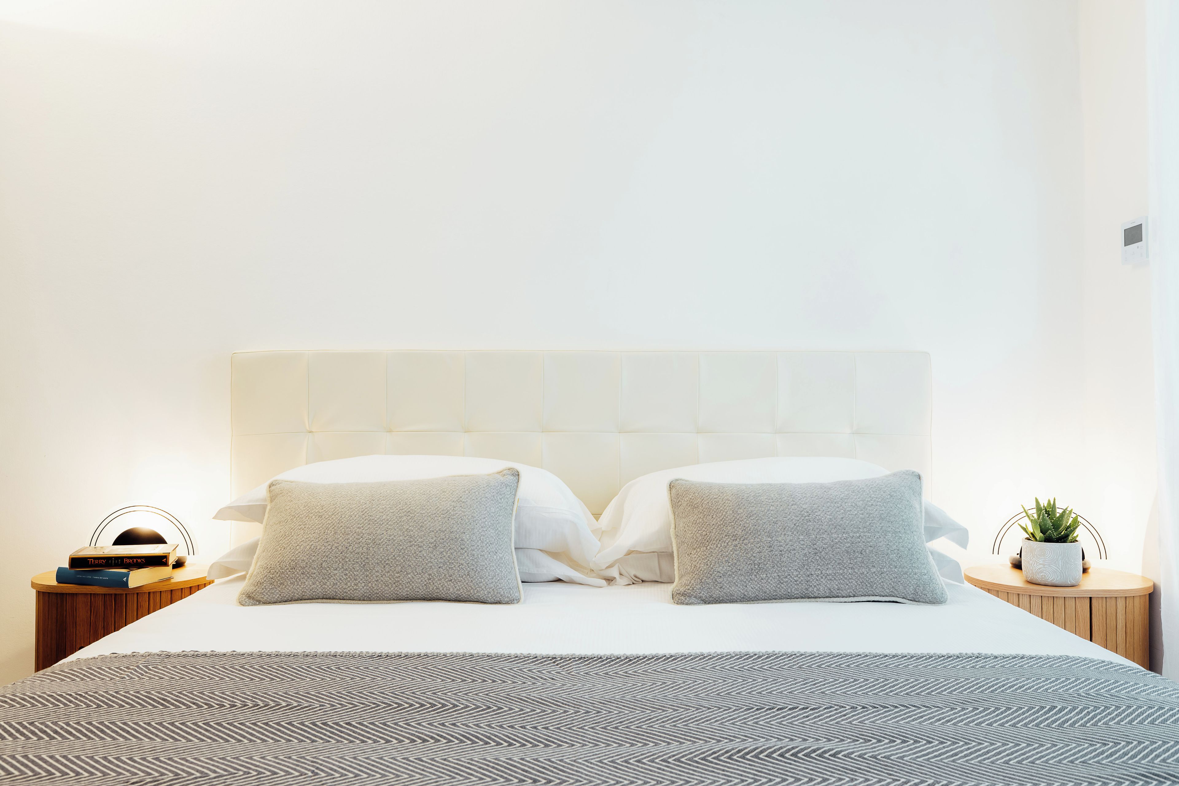 A neatly made bed with a patterned bedspread, two pillows, a bedside table with a book and a plant, and a wall-mounted thermostat.