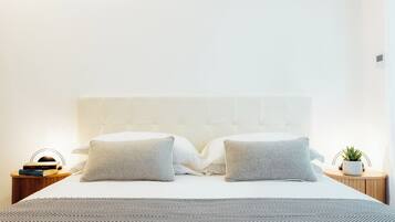 A neatly made bed with a patterned bedspread, two pillows, a bedside table with a book and a plant, and a wall-mounted thermostat.