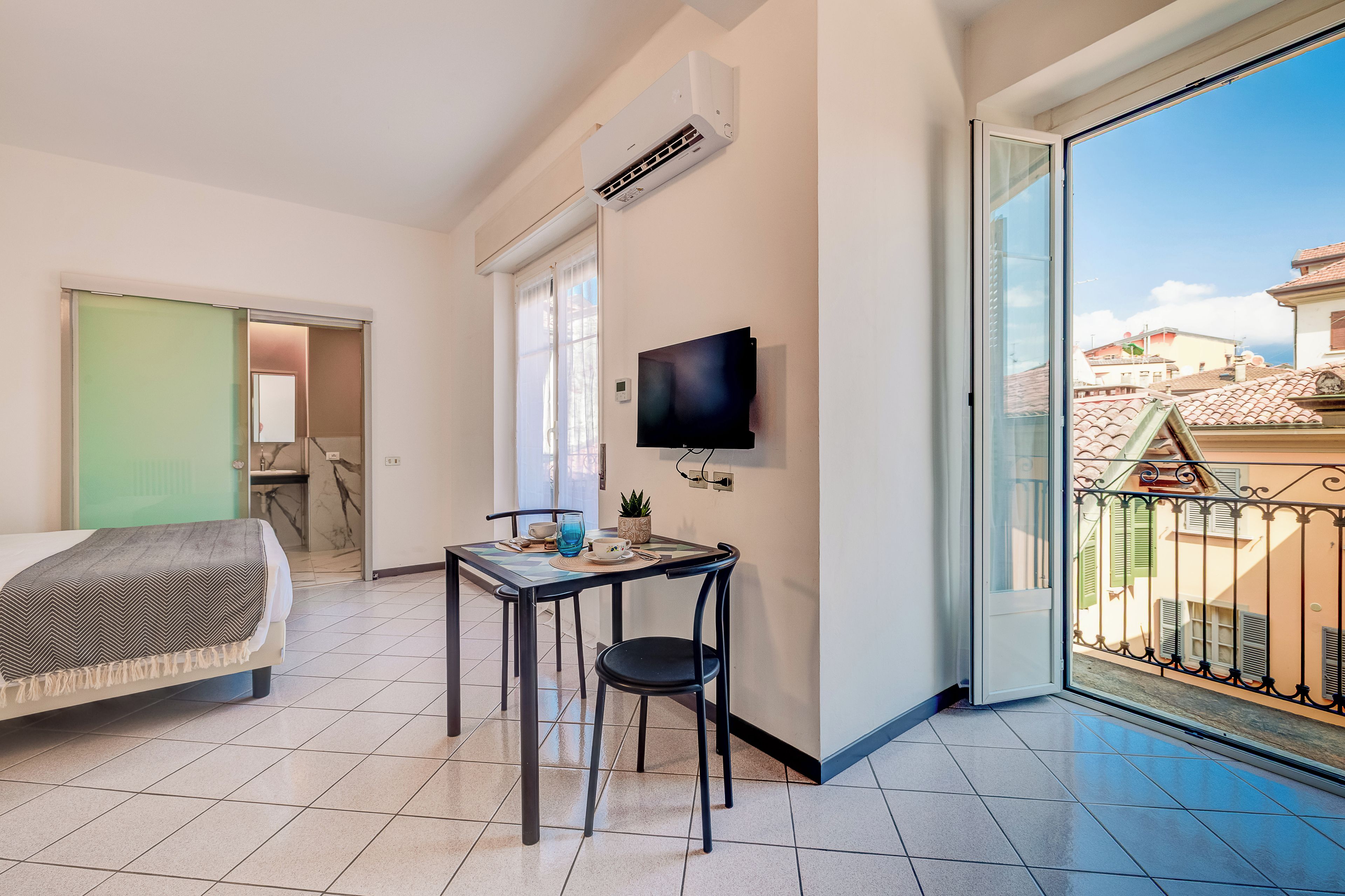 Ein Hotelzimmer mit Bett, Tisch mit Stühlen, Fernseher und Balkon mit Blick auf Gebäude.
