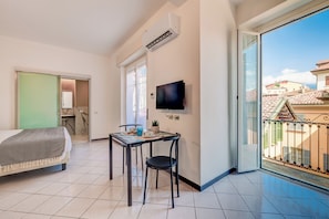 Habitación de hotel con cama, una mesa con sillas, televisión y balcón con vista a edificios.