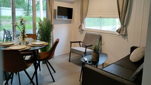 A modern room with a dining table set, a sofa, a TV mounted on the wall, and large windows with curtains. - Tiny Cottage 2+2 (Kaatsheuvel)