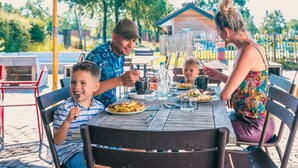 A family dining outdoors at a wooden table with chairs, a metal umbrella, and a house in the background. - Tiny Cottage 2+2 (Kaatsheuvel)