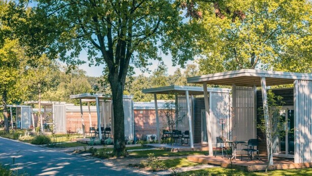 A row of mobile homes with outdoor seating areas, a paved path, and trees in the background. - Tiny Cottage 2+2 (Kaatsheuvel)