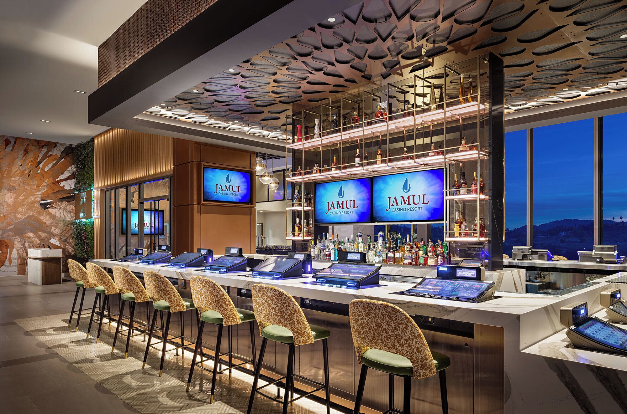 A bar area with multiple screens displaying 'Jamul Casino Resort' branding, a variety of liquor bottles, and a row of uniquely designed chairs.