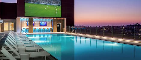 Un espace piscine avec des chaises longues, un bar et un grand écran diffusant un match de football.