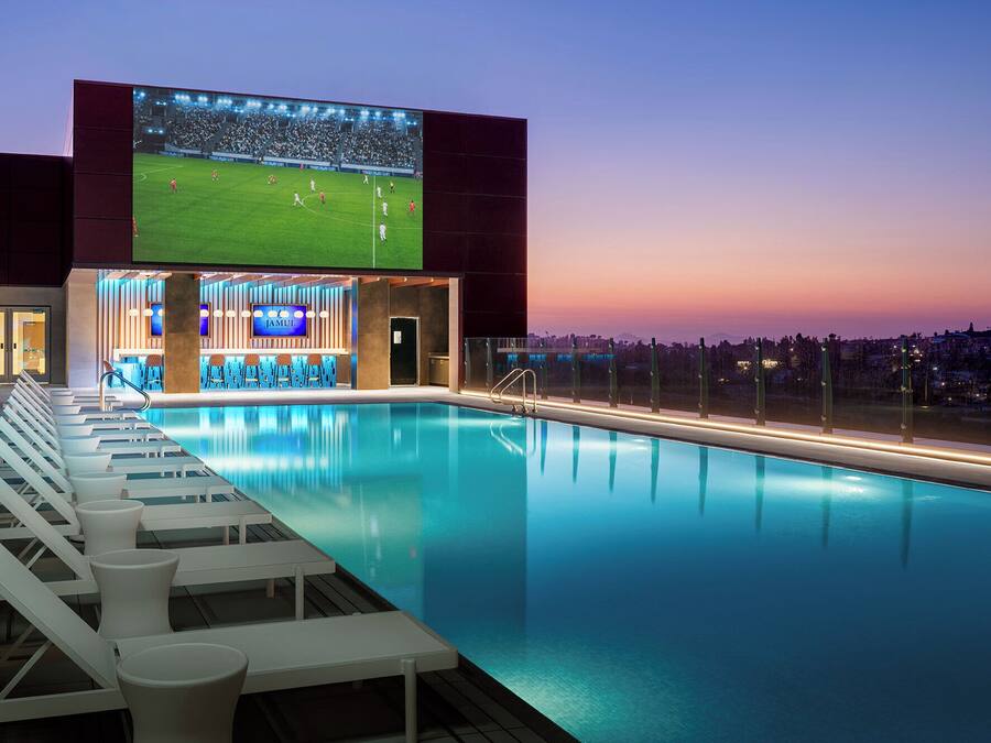 A pool area with lounge chairs, a bar, and a large screen displaying a soccer game.