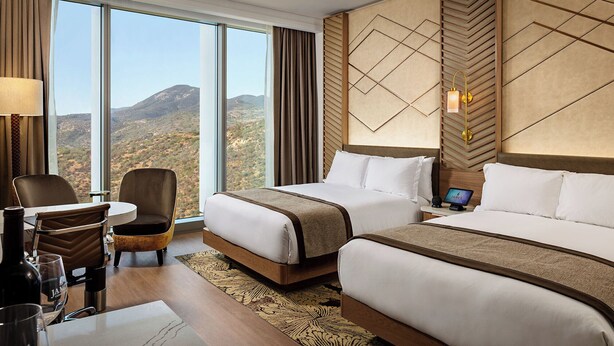Habitación de hotel con dos camas, un escritorio con silla, una lámpara y vistas a las montañas.