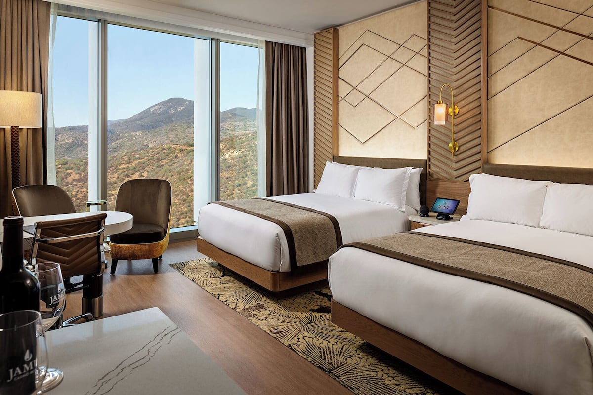 Habitación de hotel con dos camas, un escritorio con silla, una lámpara y vistas a las montañas.