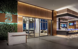 L’extérieur d’un hôtel moderne, avec un bar, un espace salon doté de tables et de chaises, et une grande enseigne lumineuse.