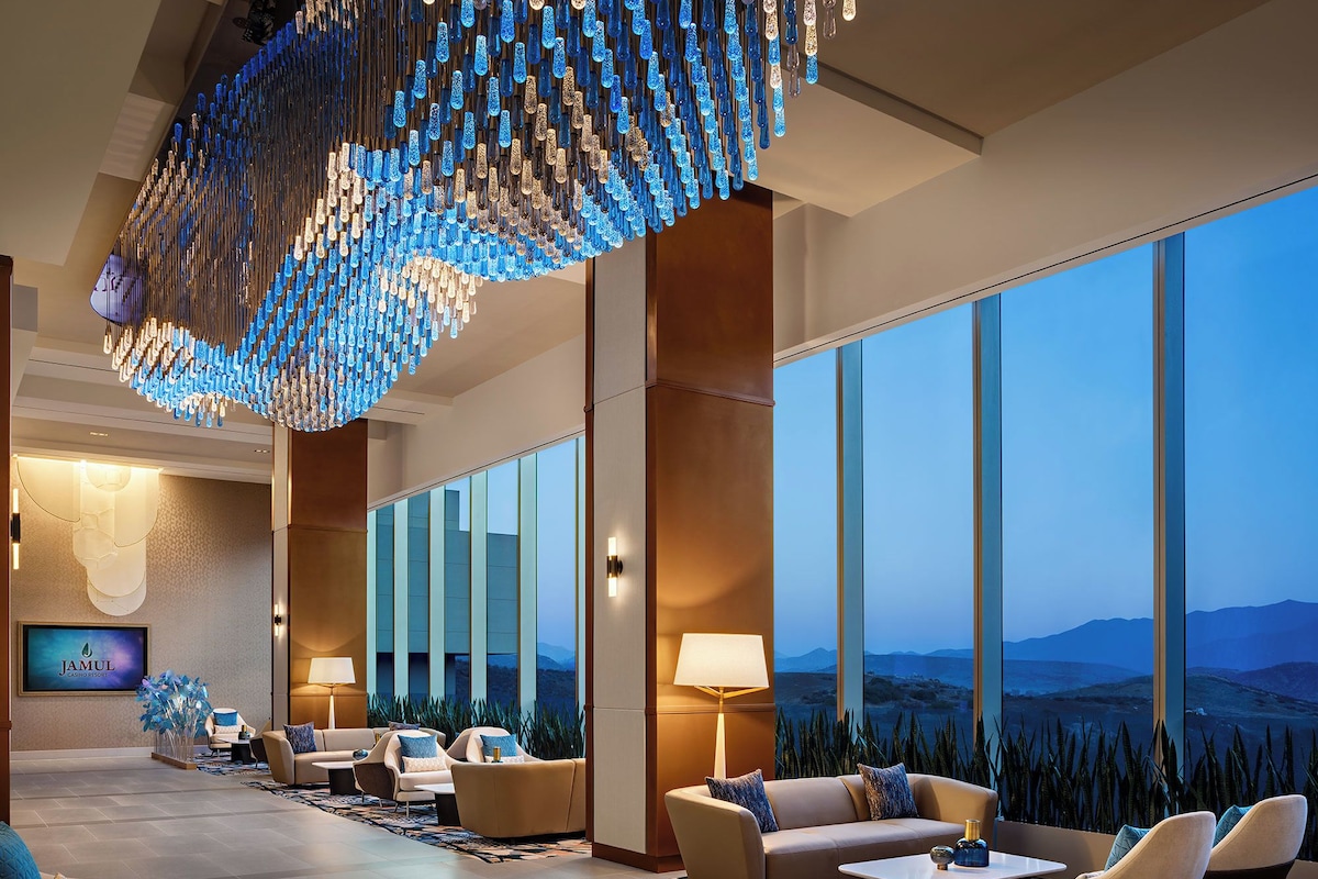 Um hall de hotel com um lustre moderno, assentos confortáveis, uma grande janela com vista para as montanhas e uma tela de televisão.