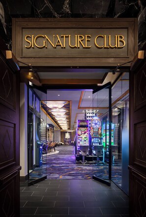 Placa 'Signature Club' acima de uma entrada com porta de vidro, levando a uma área de jogos com máquinas caça-níqueis e uma roleta.