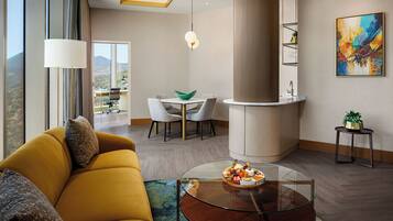 Ein modernes Wohnzimmer mit einer Couch, einem Glastisch mit Snacks, einer Essecke mit Stühlen, einer Kücheninsel und einem großen Fenster mit Blick.