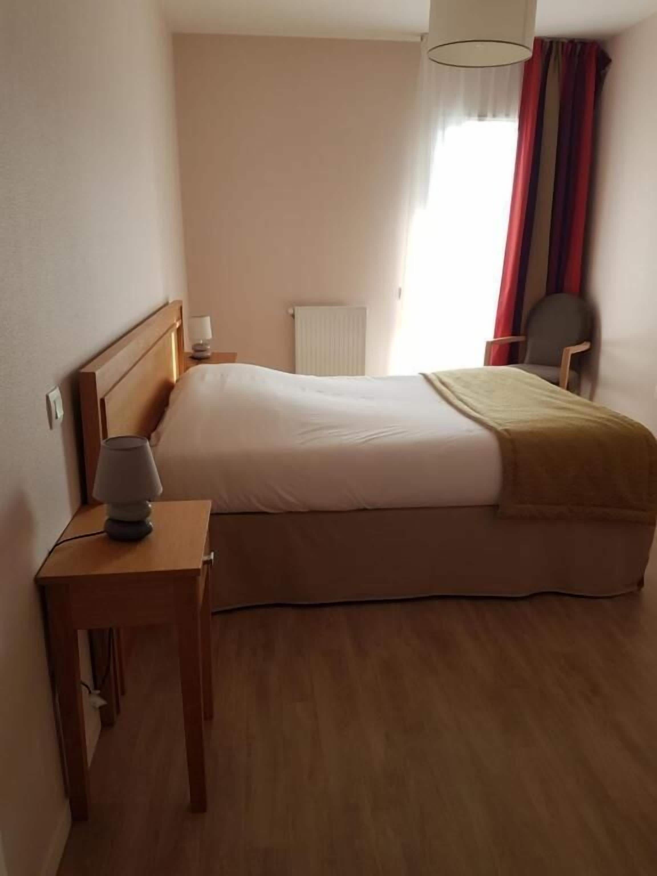 A hotel room with a bed, a nightstand, a lamp, a chair, and a window with curtains.