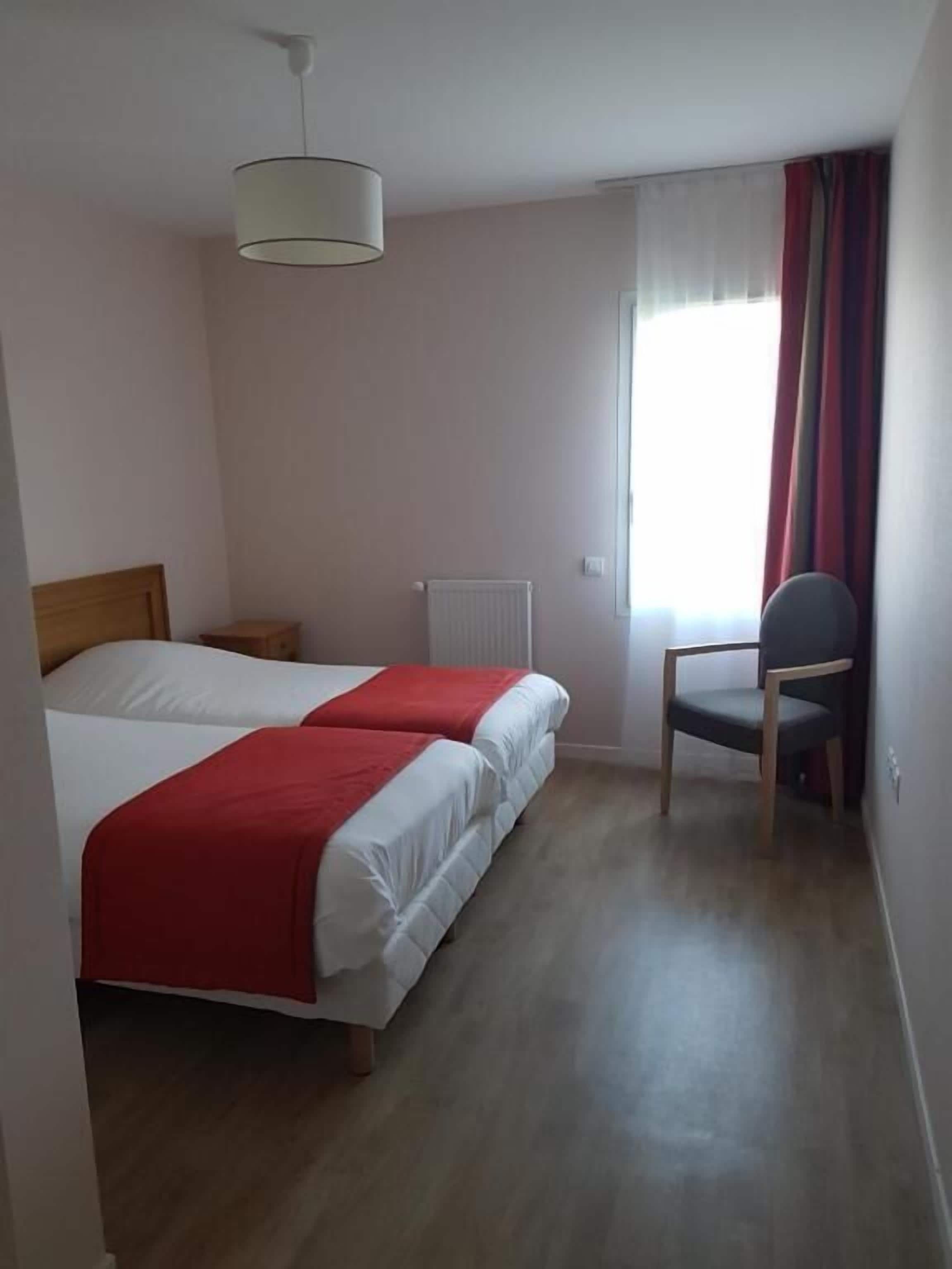 Ein Hotelzimmer mit zwei Betten, einem Sessel, einem Heizkörper und einem Fenster mit Vorhängen.