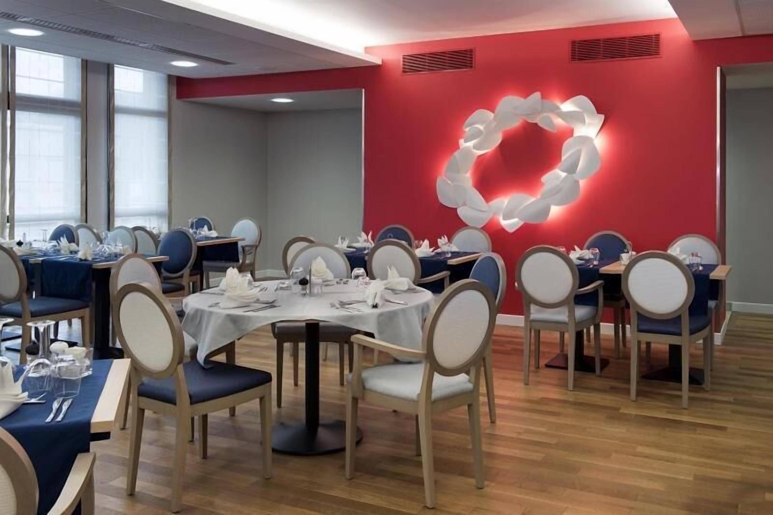 Un espace repas avec des tables et des chaises rondes, un mur d’accent rouge et un luminaire décoratif.