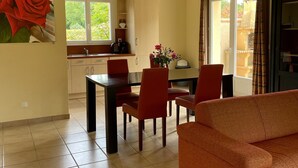 A dining area with a table and chairs, a kitchen in the background, and a large window with curtains.