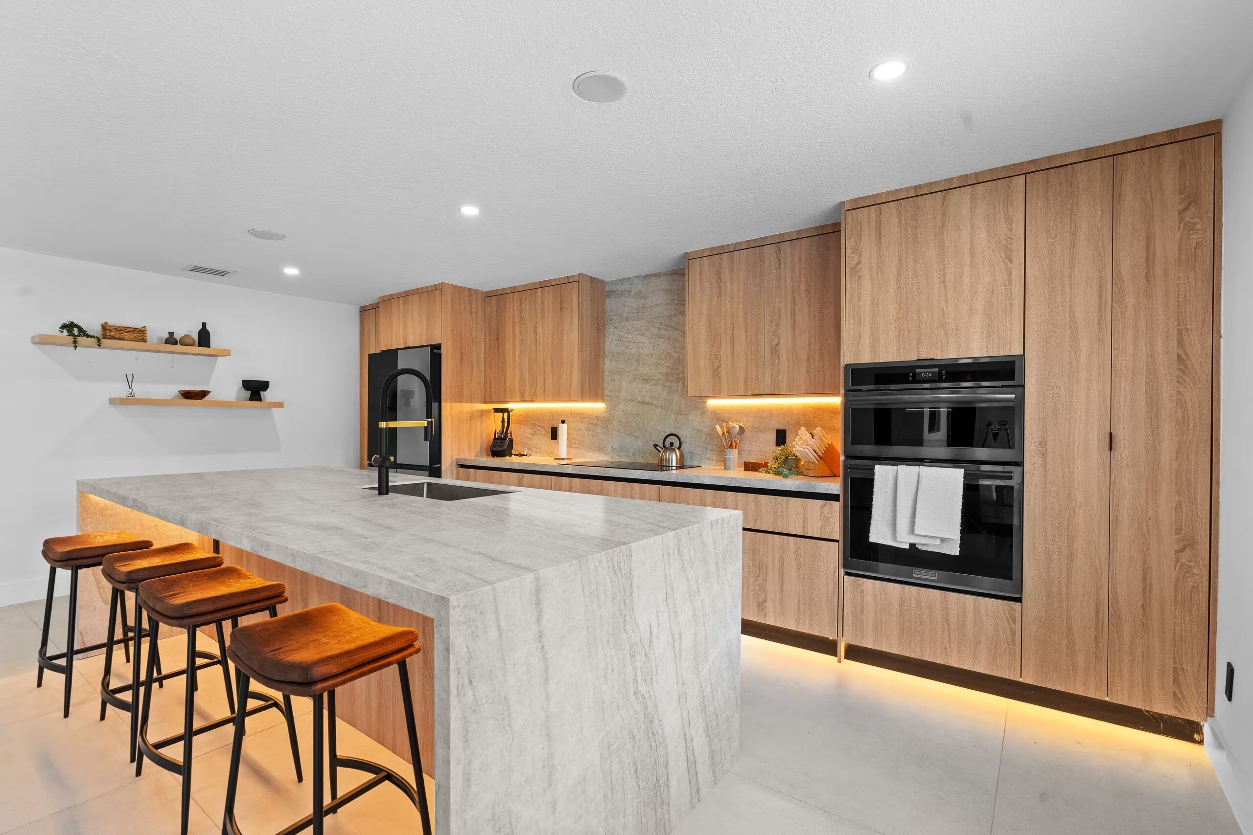 A modern kitchen with wooden cabinets, a marble island, bar stools, a refrigerator, an oven, and under-cabinet lighting.
