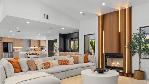 Ein modernes Wohnzimmer mit einem Ecksofa, einem runden Couchtisch, einem Kamin und einer Küche im Hintergrund.