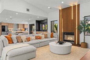 Een moderne woonkamer met een hoekbank, een ronde salontafel, een open haard en een keuken op de achtergrond.