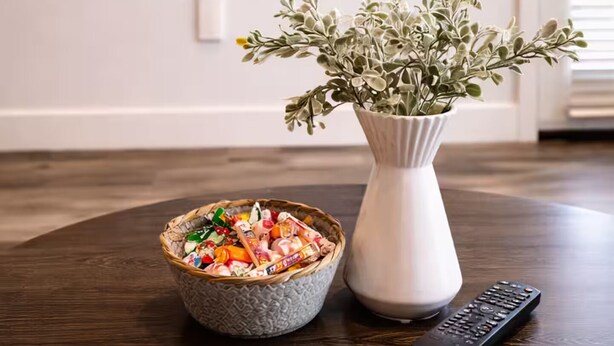 Et værelse med et fjernsyn ophængt på væggen, en vase med en plante, en skål med snacks og en fjernbetjening på et træbord.