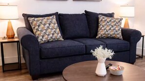 En mørkeblå sofa med mønstrede puder, en hvid vase med blomster og en fjernbetjening på et rundt træbord. Der er to lamper på hver side af sofaen.