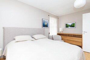 A hotel room with a large bed, two pillows, a bedside table with a lamp, a chair, and two framed pictures on the wall.