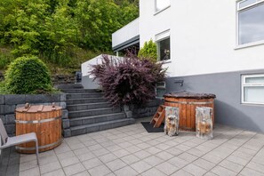 A modern outdoor patio with a wooden hot tub, a bench, and a staircase leading to a building.