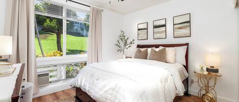 A bedroom with a bed, a ceiling fan, a window with curtains, a bedside table with a lamp, and a wall-mounted artwork.