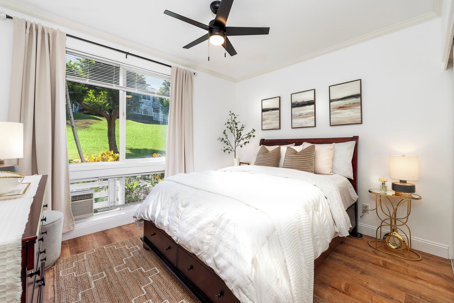 Quarto com cama, ventoinha no teto, janela com cortinas, mesa de cabeceira com abajur e uma obra de arte na parede.