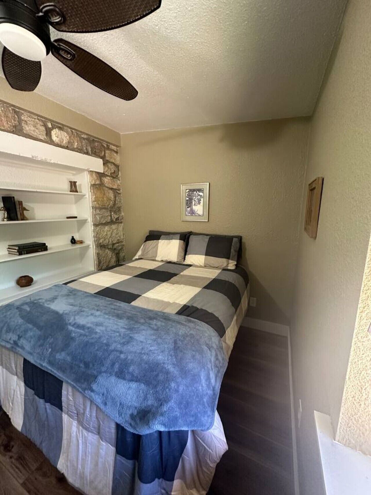 Un dormitorio con cama, ventilador de techo, pared de piedra, estante con libros e imagen enmarcada en la pared.