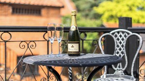 A bottle of champagne on a metal table with two glasses, set on a balcony with a wrought iron railing and a brick building in the background.