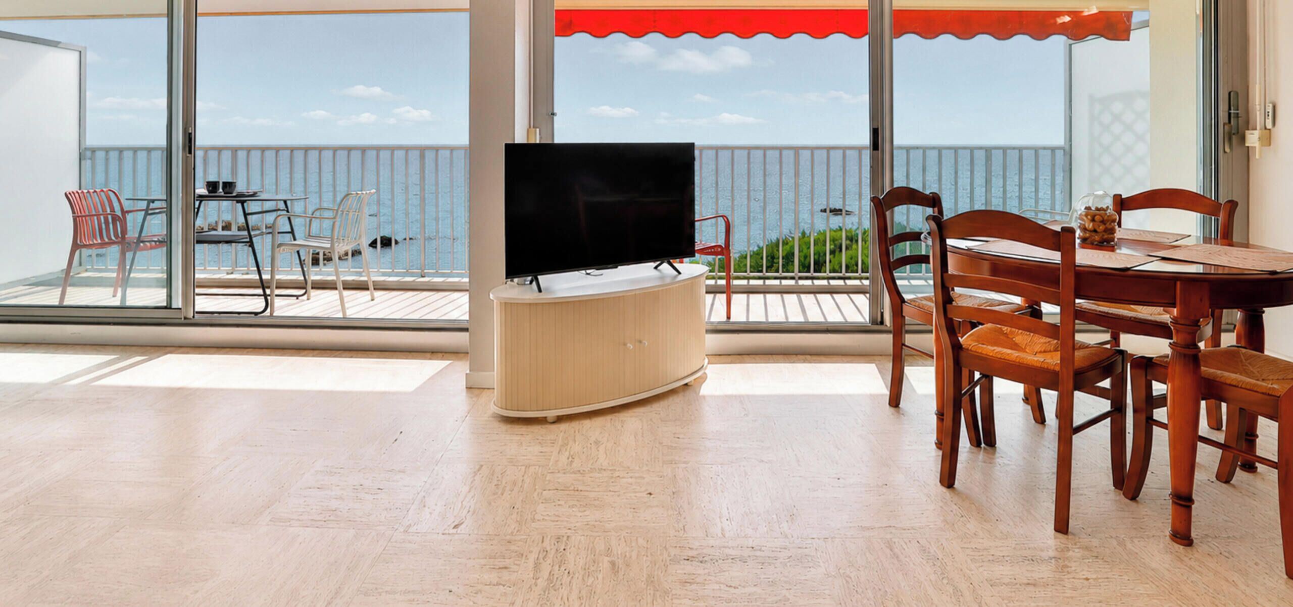 A balcony with a TV, dining table, chairs, and a view of the sea.