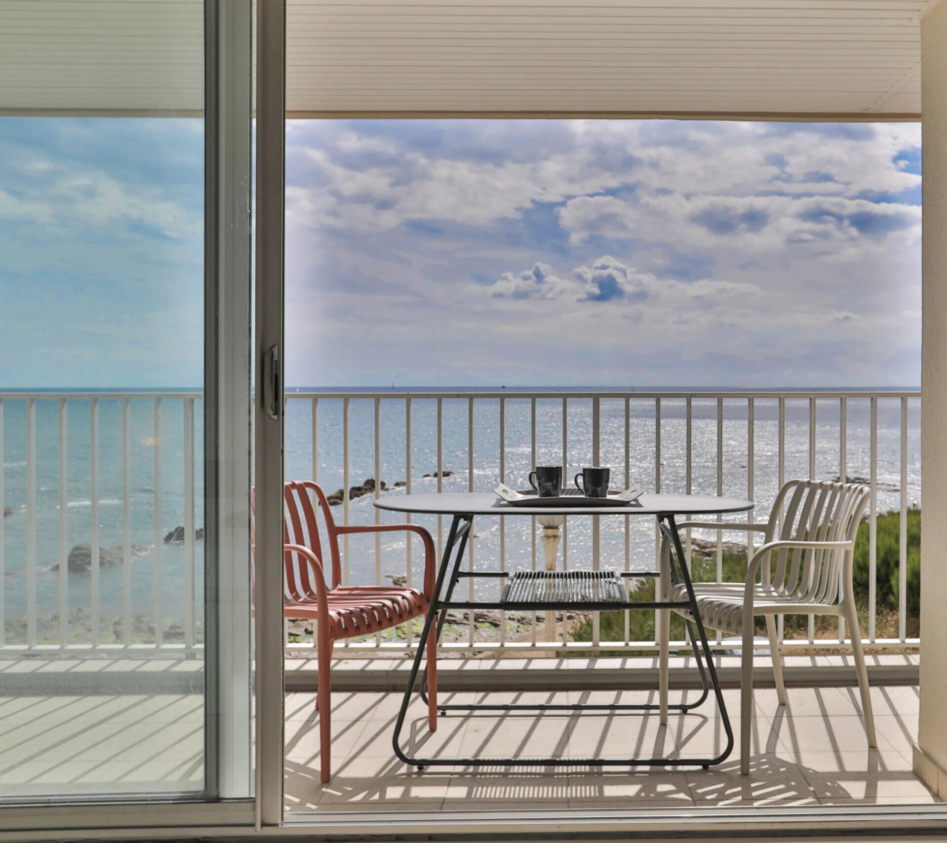 Ein Balkon mit Tisch und Stühlen, der Blick auf einen Strand unter blauem Himmel bietet.