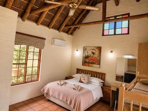A bedroom with a wooden ceiling, a bed with a pink bedspread, a desk, and a window with a view of greenery. - Stonebreaker Country Lodge  (Oudtshoorn)