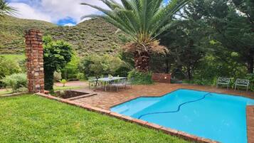 Piscina com borda de tijolos, cercada por grama e árvores, com uma estrutura de tijolos e uma mesa com cadeiras por perto.