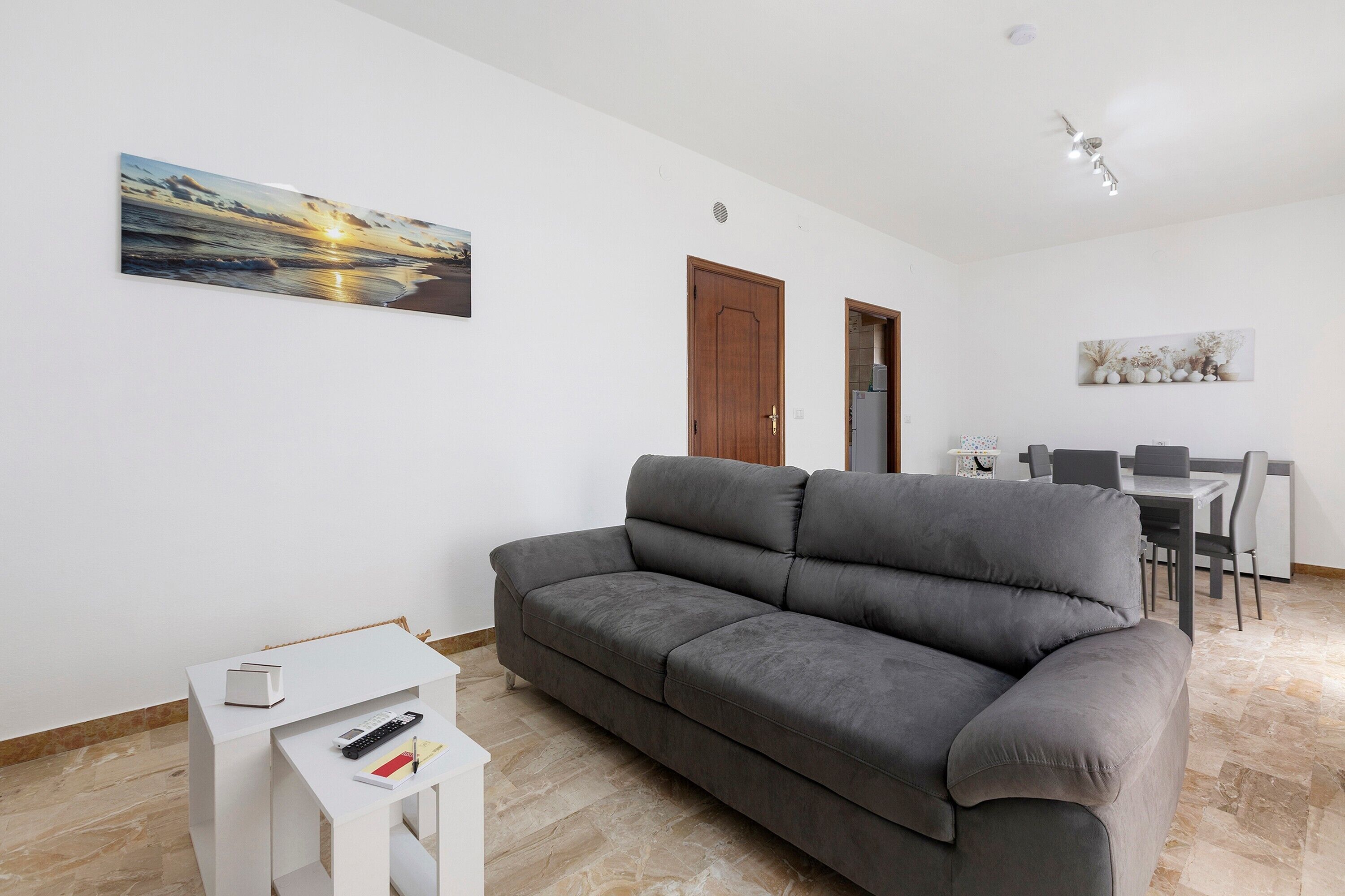 Un salon avec un canapé gris, une petite table blanche sur laquelle se trouve une télécommande, et un tableau accroché au mur.