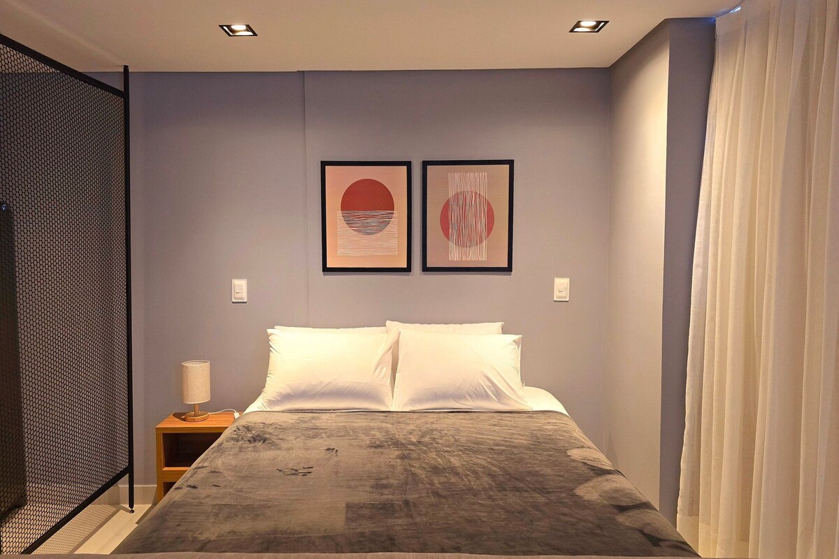 Ein Schlafzimmer mit einem Bett, zwei an der Wand befestigten Kunstwerken, einem Nachttisch und einem Fenster mit Vorhängen.