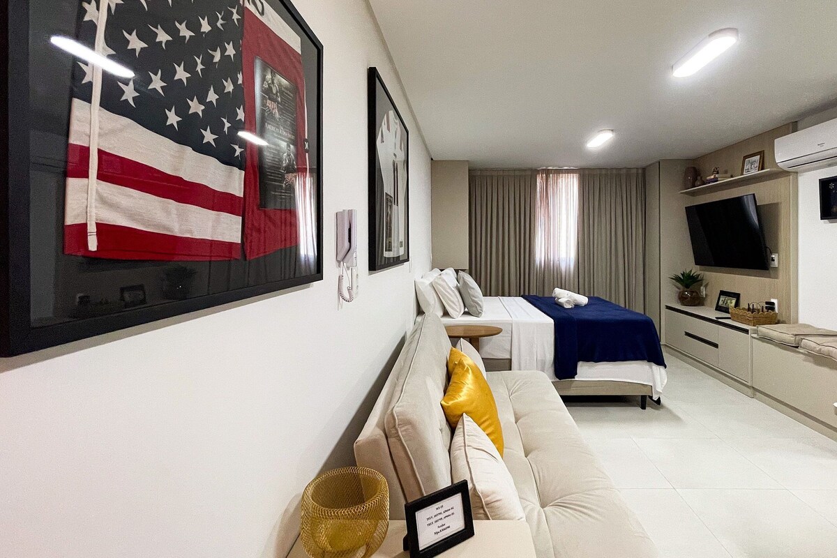 Ein Hotelzimmer mit einer großen US-amerikanischen Flagge, einem Bett mit blauer Bettdecke, einem Sofa, einem kleinen Tisch und einem Fernseher an der Wand.