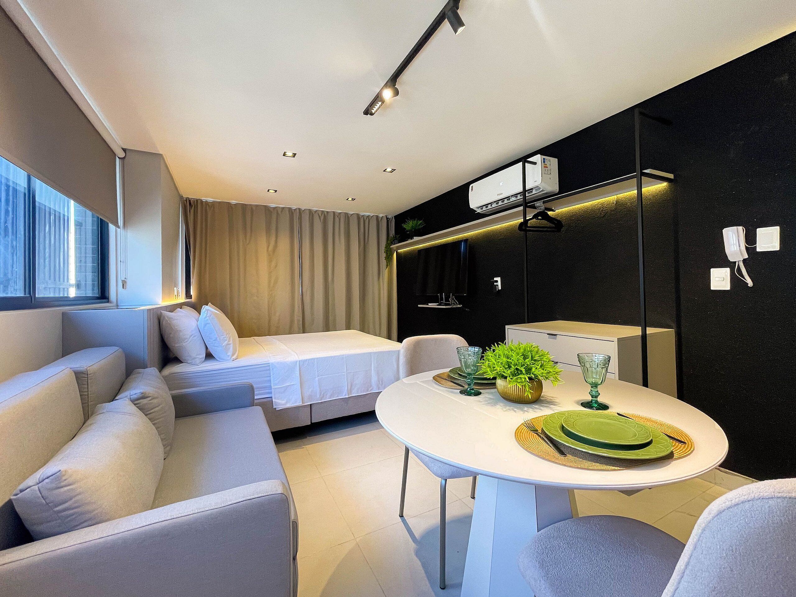 A modern hotel room with a sofa, a small bed, a dining table with green plates, and a potted plant.