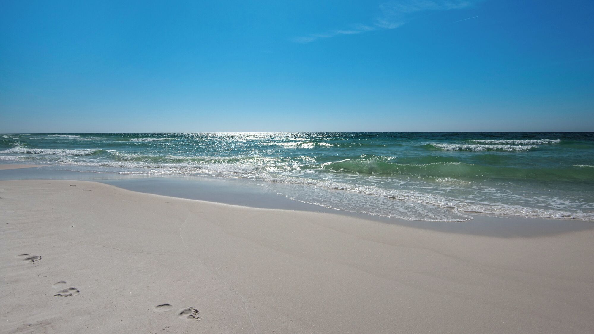 A beach with footprints leading to the water, a clear sky, and waves approaching the shore.