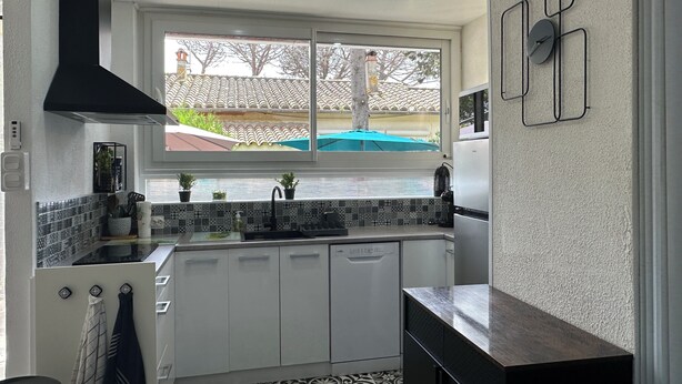 Una cuina moderna amb armaris blancs, una campana extractora negra i una finestra amb vista a una casa i arbres.