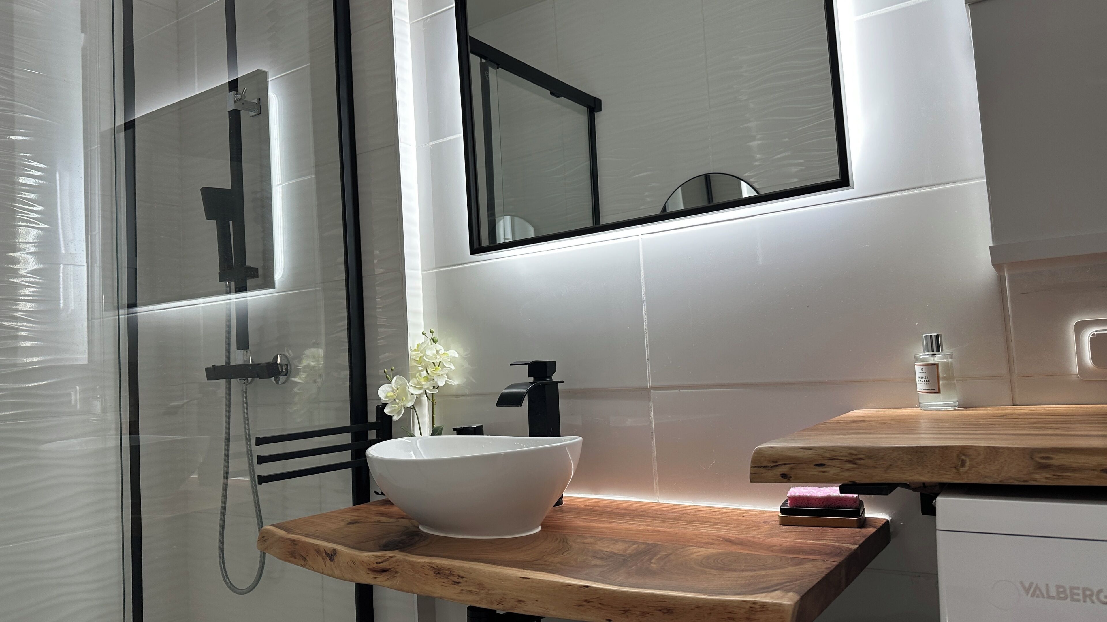 Une salle de bain moderne avec une douche à parois de verre, un lavabo blanc doté d’un robinet noir et un plan de toilette en bois.